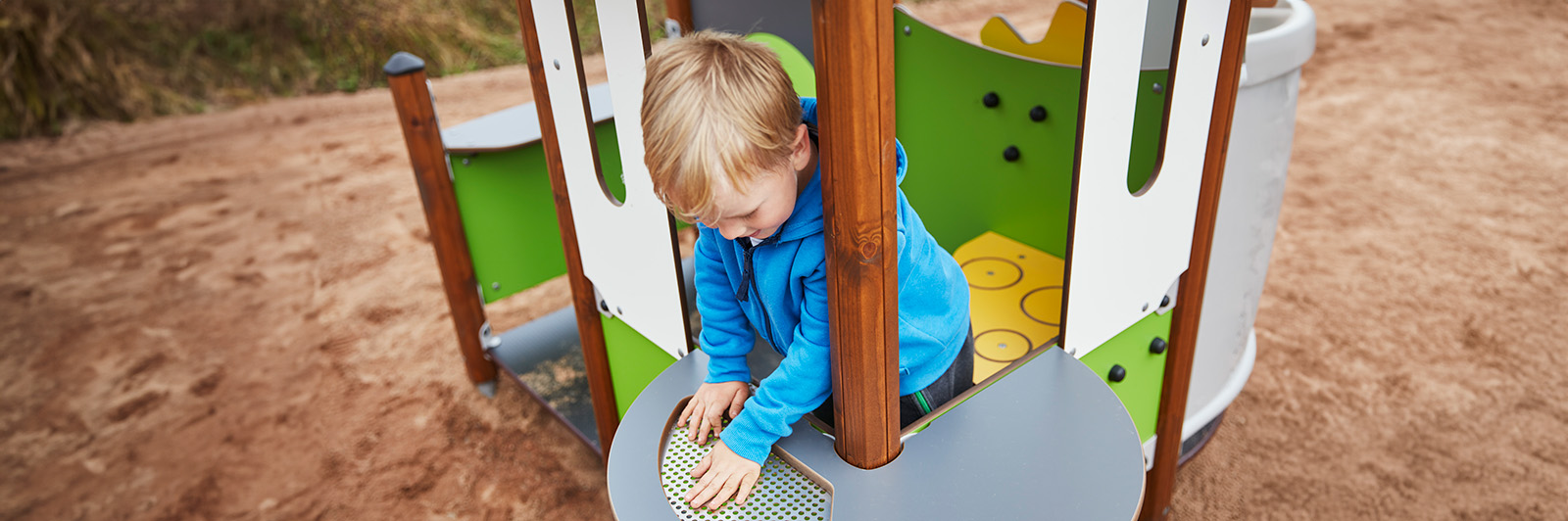 Ein Junge interagiert mit Spieltafeln auf einem Spielplatz, in den ein Spielhaus eingebaut ist.